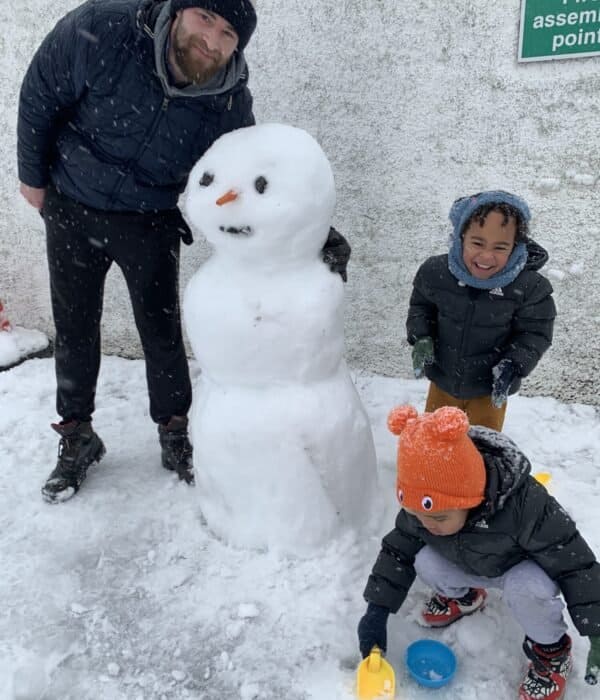 Family posing with a snowman in the Irish countryside during Christmas, smiling and bundled up in winter clothing.