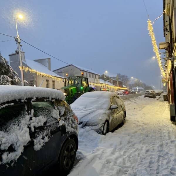 A tractor driving through a snow-covered town in the Irish countryside, surrounded by frost-laden cars and houses.